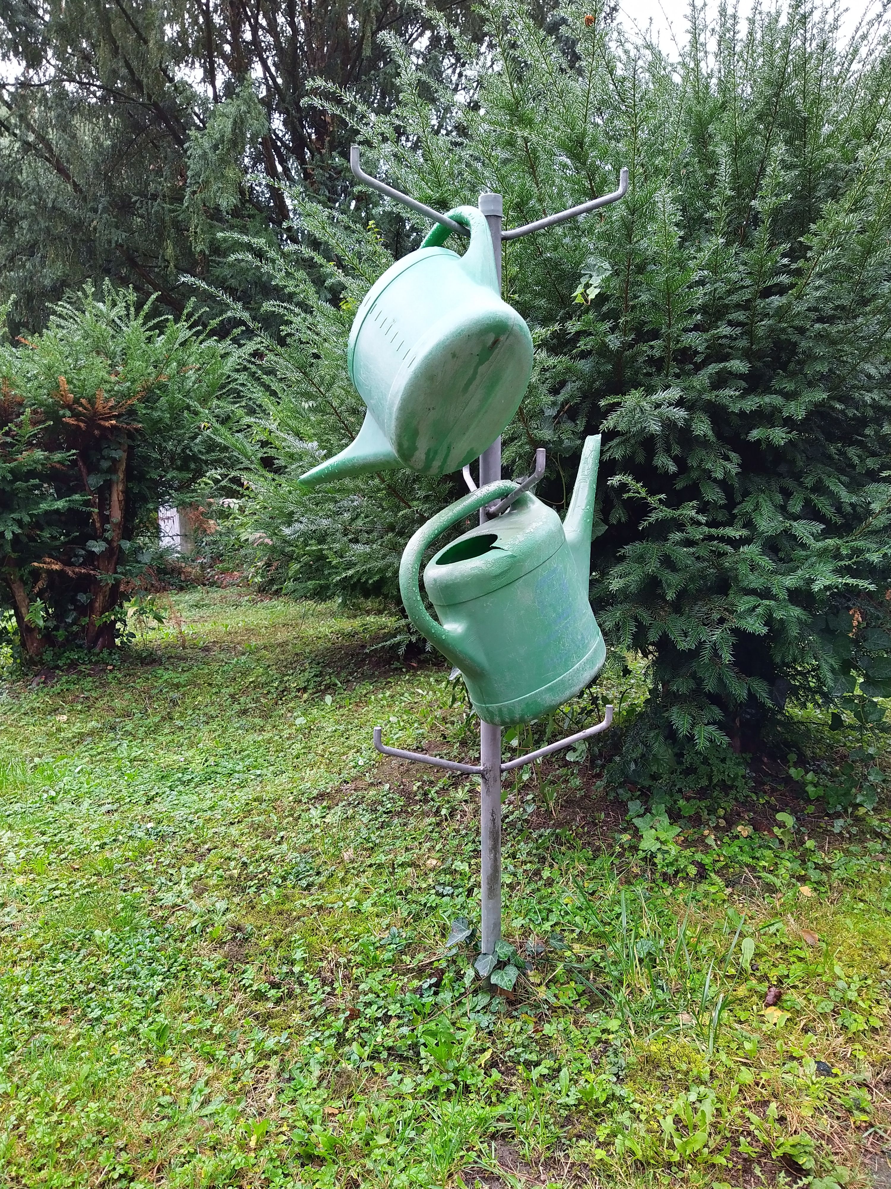 Grüne Gießkannen an einem Halter zwischen den Gehölzen des Friedhofs Höchst